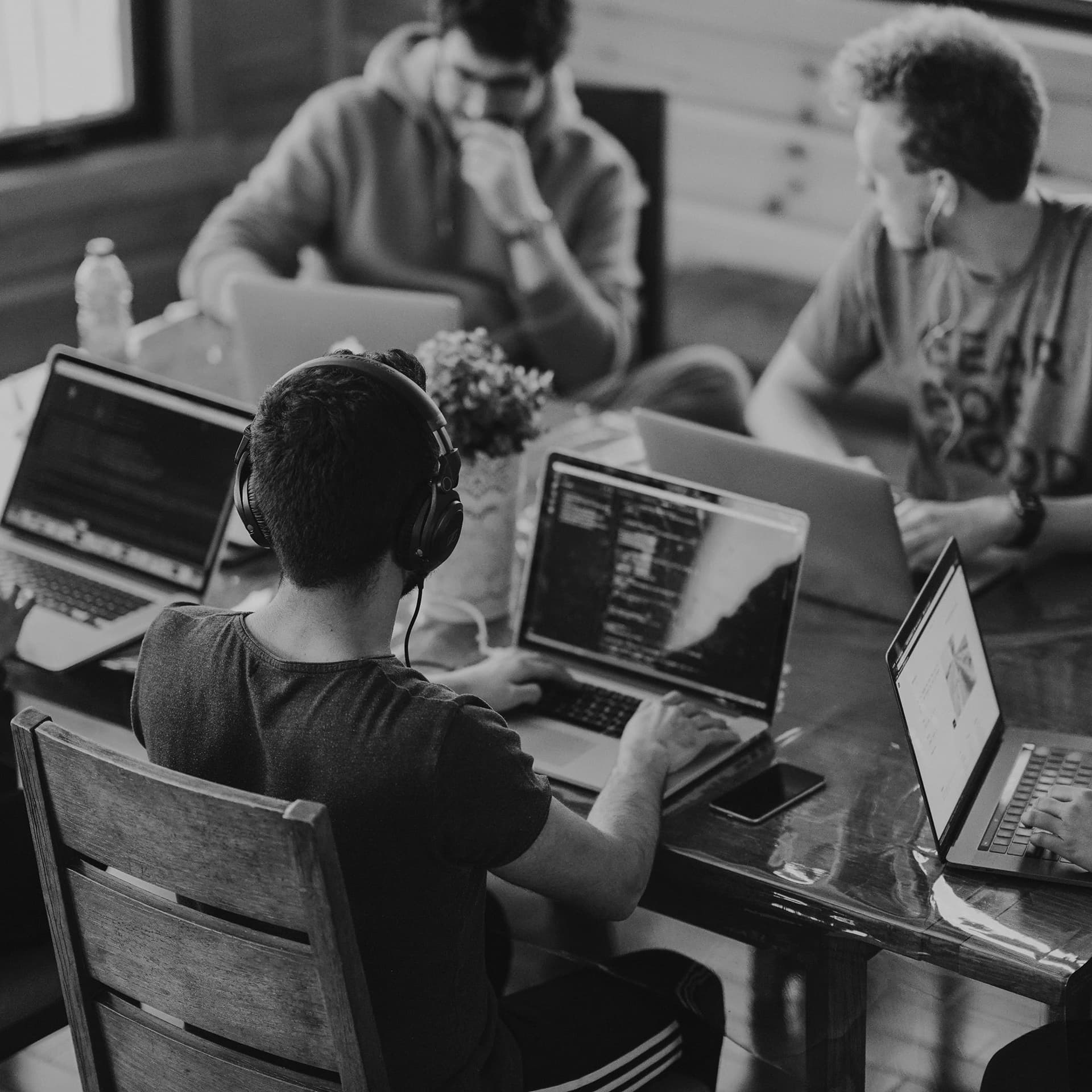 Black and white photo of four people working on laptops around a table. One person in the foreground wears headphones.
