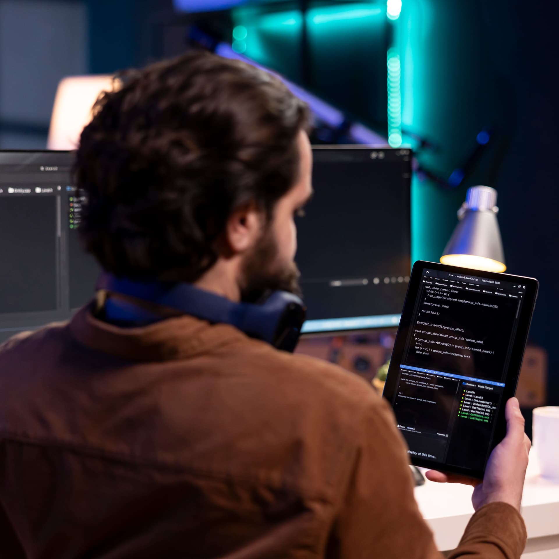 Man from behind viewing code on a tablet and a computer monitor in a dark office with neon lighting.
